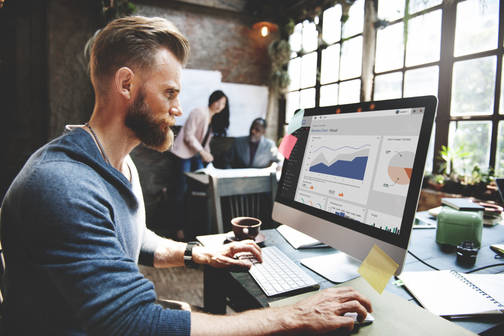 Man with beard looking at a graph in the foreground while two office workers work in the background